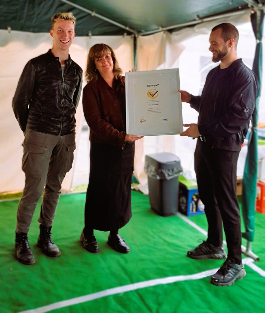 Das Bild zeigt drei Menschen, zwei halten ein Zertifikat in einem Bilderrahmen